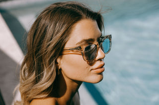 Woman by a swimming pool wears peach clear biodegradable and sustainable ike oat clue sunglasses with blue lens